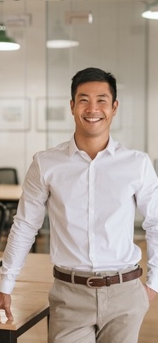 Smiling young Asian businessman standing next to a table while working alone in a large modern office